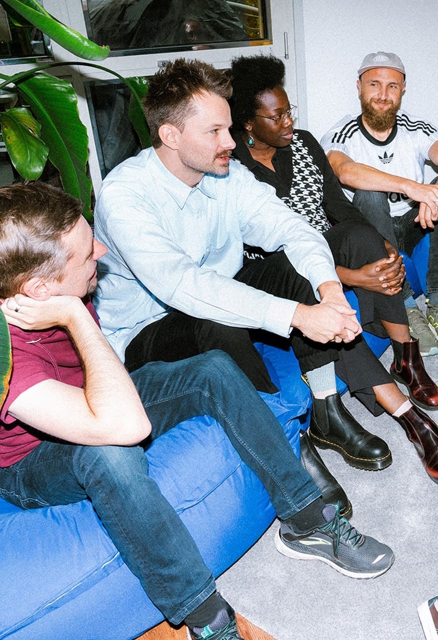 Four young adults are sitting in a circle on blue beanbags in front of a wooden slatted wall, laughing.