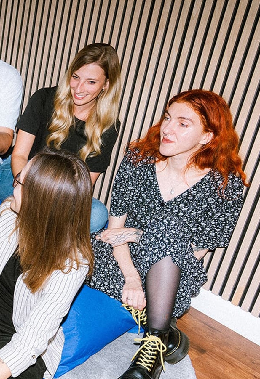 Four young adults are sitting in a circle on blue beanbags in front of a wooden slatted wall, laughing.